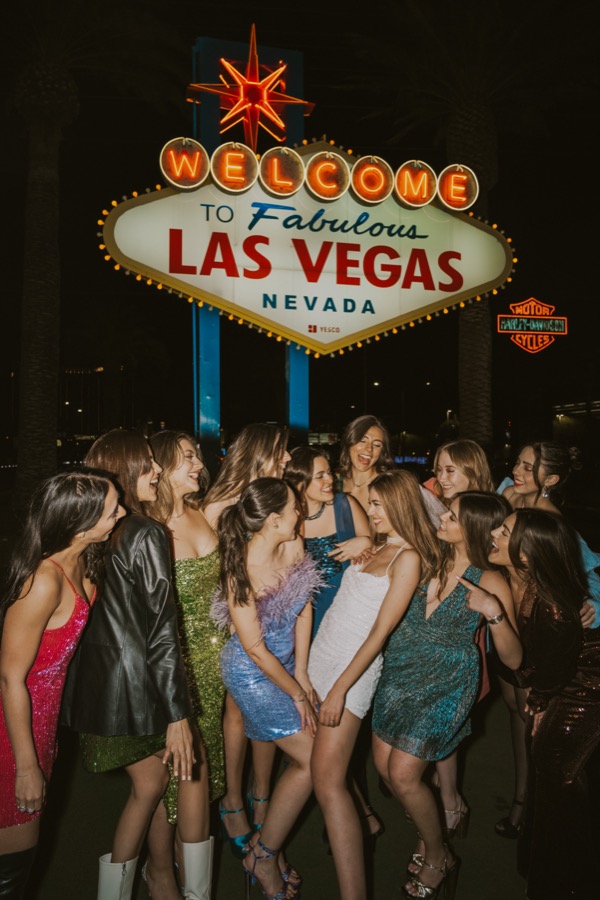 A group of friends in sparkly dresses in front of the Welcome to Las Vegas sign at night